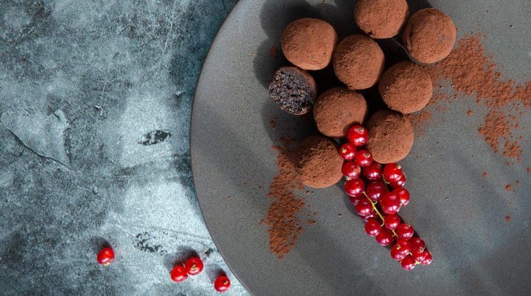 Mandelmehltrüffel mit Kakao bestäubt auf braunem Teller mit roten Johannisbeeren von oben.