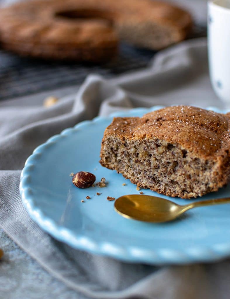 Nahaufnahme eines Stücks Haselnusskuchen auf blauem Teller mit goldfarbenem Löffel, im Hintergrund unscharf der restliche Kuchen.