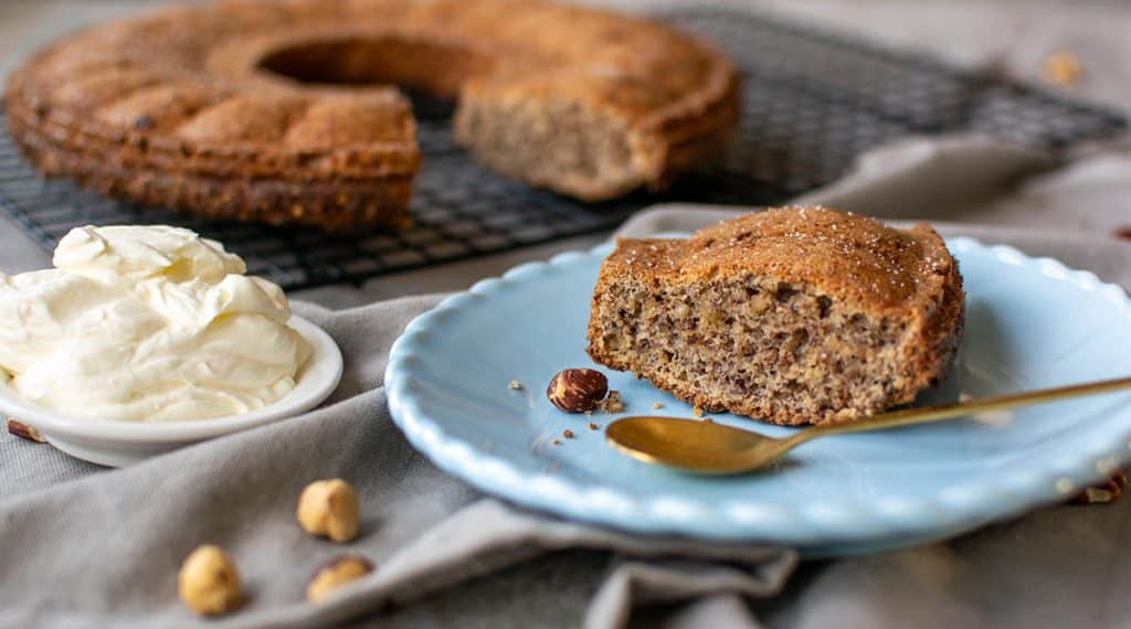 Stück Nusskuchen auf blauem Teller mit Löffel, daneben Haselnüsse und eine Schale mit Sahne.