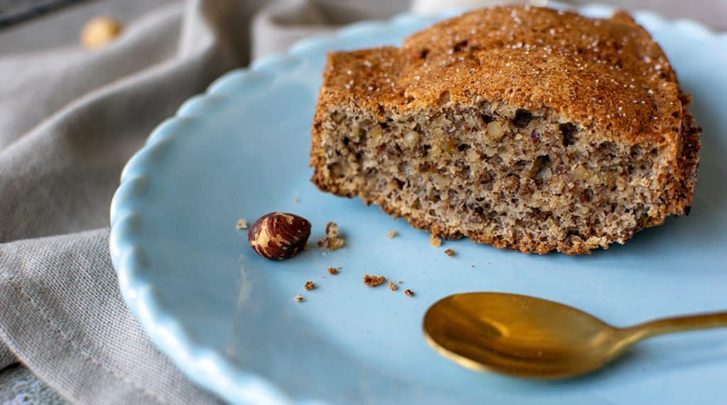 Stück Nusskuchen auf einem hellblauen Teller mit goldfarbenem Löffel, daneben eine Haselnuss und Krümel.