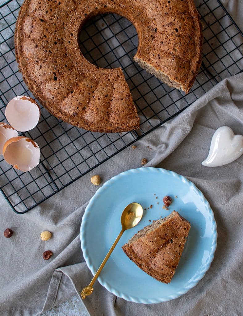 Unten ein Stück Haselnusskuchen aus der Vogelpespektive auf einem blauen Teller mit goldfarbenem Löffel. Oben der angeschnittene Kuchen mit leeren Eierschalen daneben. 