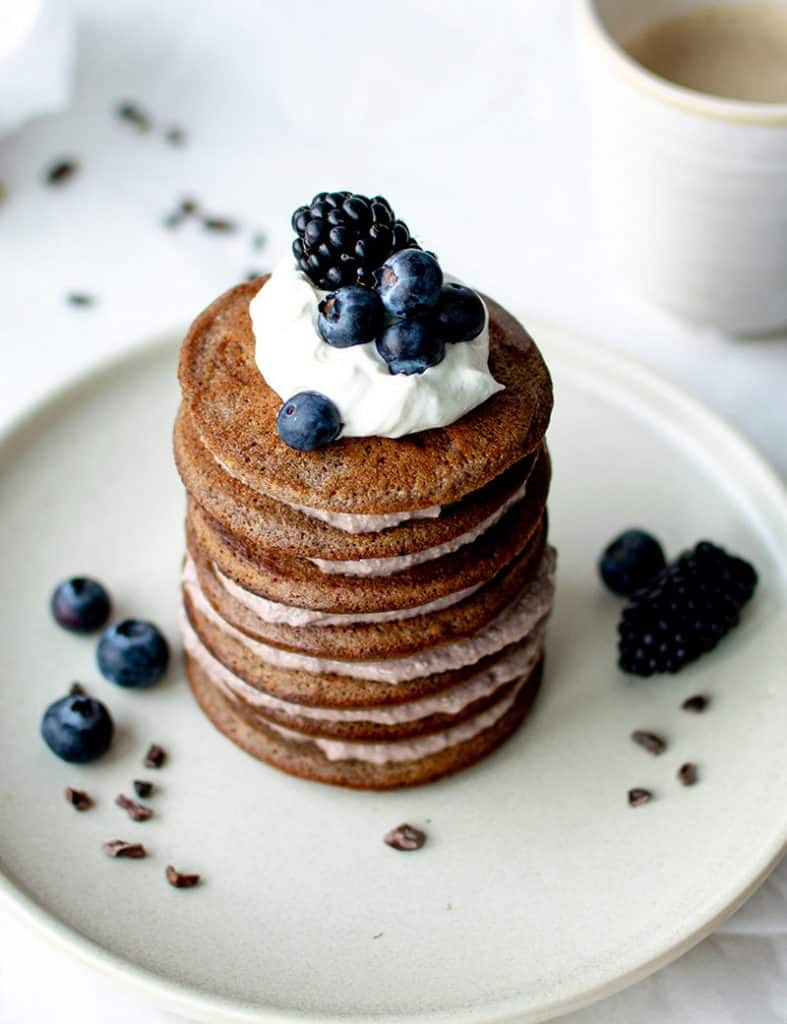 Ein Turm mit 7 Mandelmehl Pancakes, dazwischen eine Schicht mit Kakao-Quarkcreme. Oben als Deko ein paar blaue Beeren auf einem Klecks Sahne-Mascarpone-Creme. 