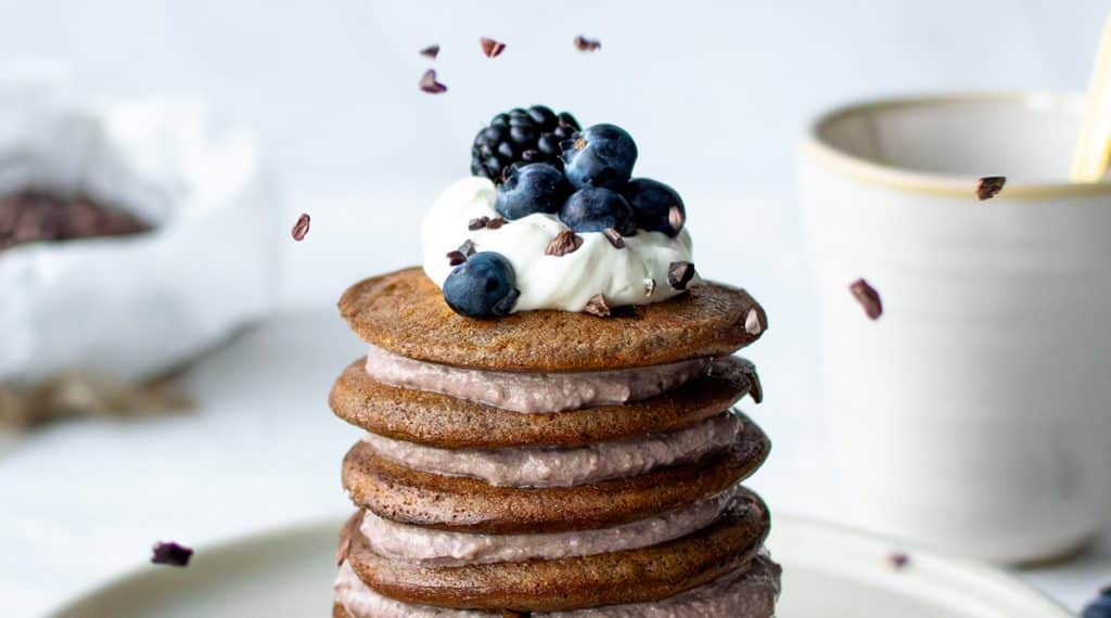 Stapel aus Mandelmehl-Pancakes mit Quarkcreme, Blaubeeren und fallenden Kakaonibs