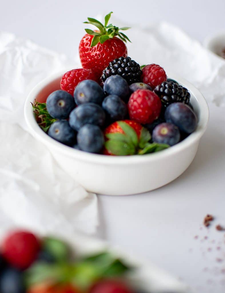Verschiedene Beeren in einer weißen Schale. Erdbeeren, Heidelbeeren, Brombeeren und Himbeeren. Eine Erdbeere lugt heraus und hat noch einen grünen Strunk. 
