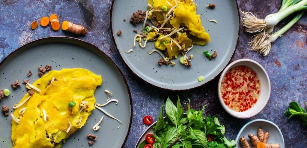 Ansicht von oben: 2 Teller mit Banh Chhaev, dazu die passende Sauce in einem Schälchen mit Fischsauce, gehackten Erdnüssen und Chilli und ein Teller mit Kräutern. 