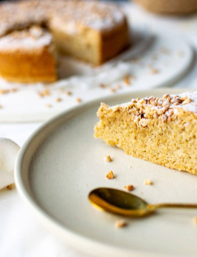 Vorn im Bild rechts die Spitze von einem Stück Mallorquinischen Mandelkuchen auf einem Teller mit einem goldenen Löffel. Im Hintergrund der angeschnittene Mallorquinische Mandelkuchen. 