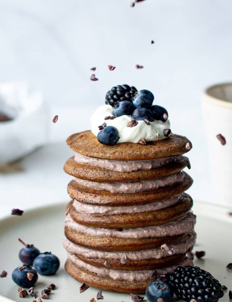 Mandelmehl-Pancakes als gestapelter Turm mit Quarkcreme, Sahne und Blaubeeren. Drumherum Kakao-Nibs, die gerade herunterfallen. 