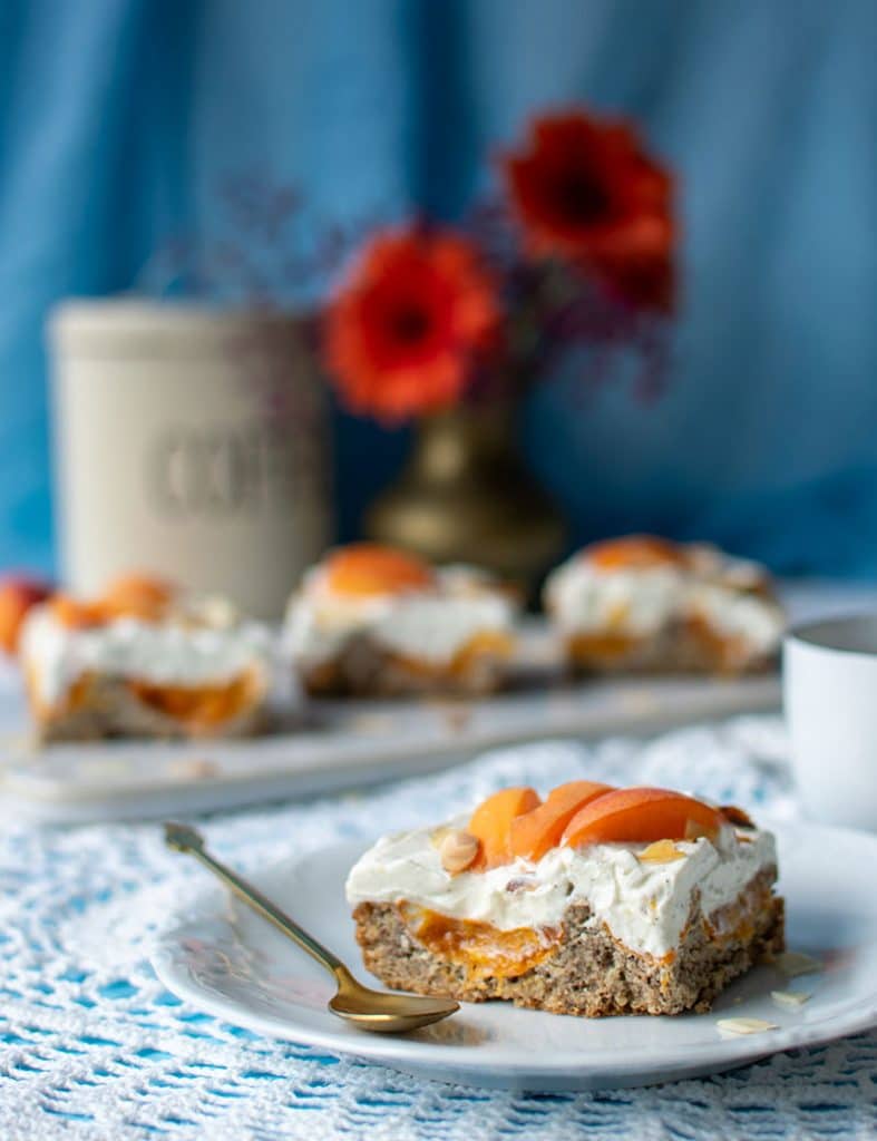 Großansicht von einem Stück Haselnusskuchen mit Aprikosen und Creme. Im Hintergrund verschwommen drei weitere Stücke vom Kuchen auf einer rechteckigen Kuchenplatte. 