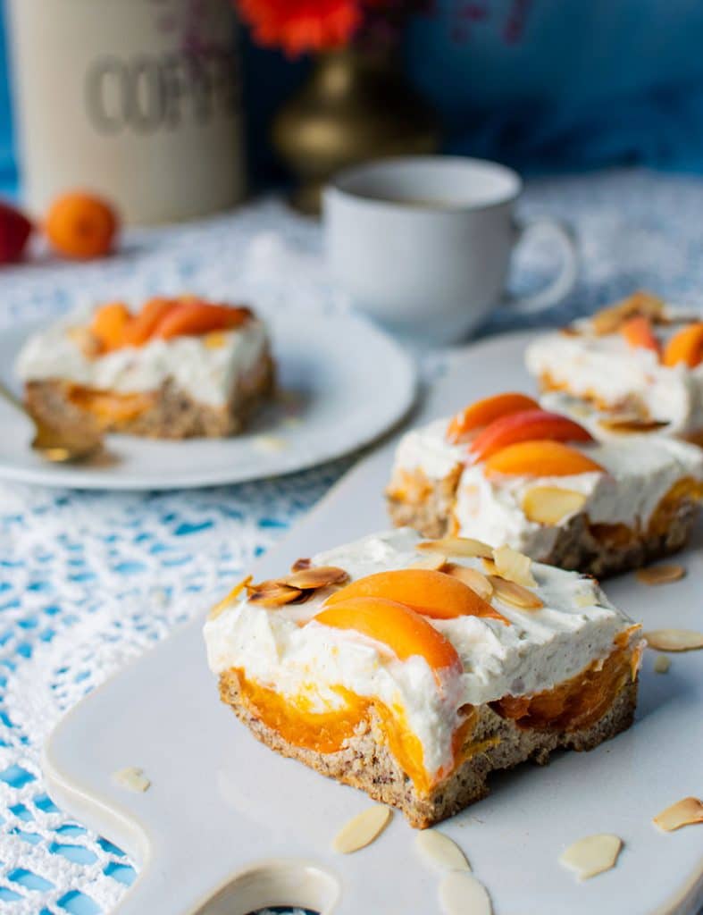 Saftiger glutenfreier Haselnusskuchen mit Aprikosen, Creme und gerösteten Mandelblättchen auf einer rechteckigen Kuchenplatte aus weißem Porzellan.
