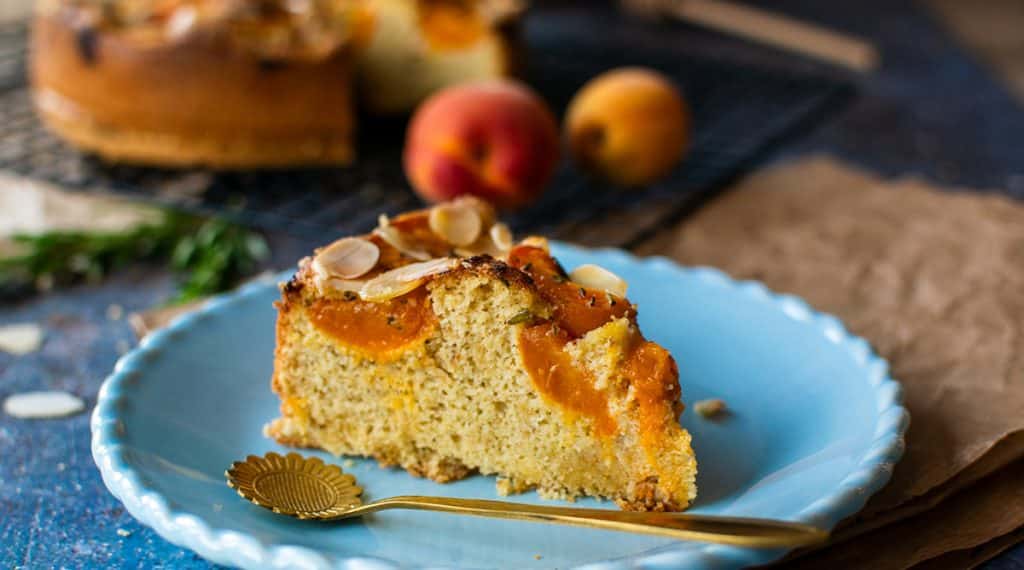 Ein Stück Mendelmehlkuchen mit einem Belag aus frischen Aprikosen auf einem hellblauen Teller mit einem goldfarbenem Löffel davor.
