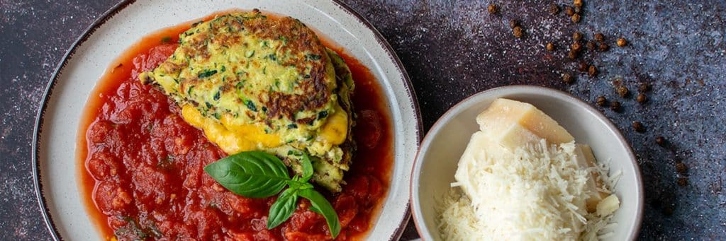Low-Carb Zucchini-Puffer mit Käse und Tomatensauce angerichtet mit frischem Basilikum. Rechts daneben eine kleine Schale mit gehobeltem Parmesan.