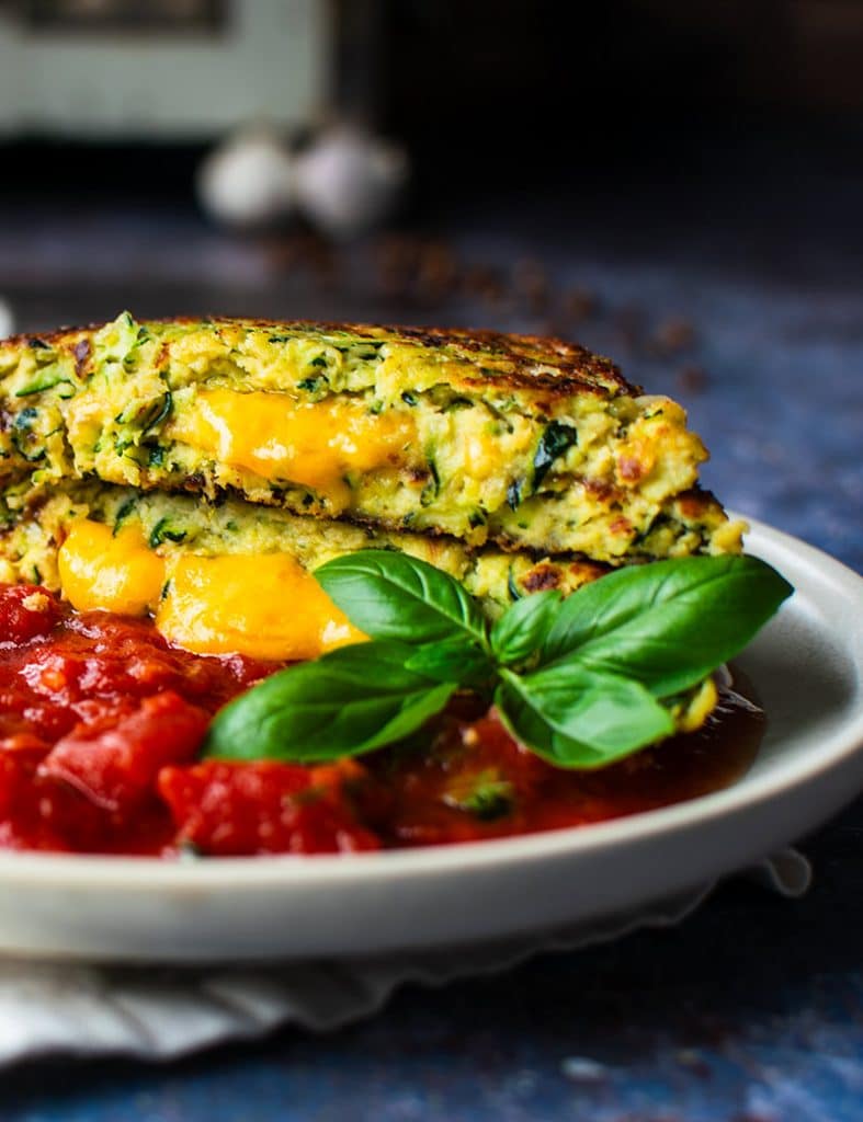 Aufgeschnittene Zucchini-Puffer mit cremigem Käsekern und fruchtiger Tomatensauce von vorn fotografiert.